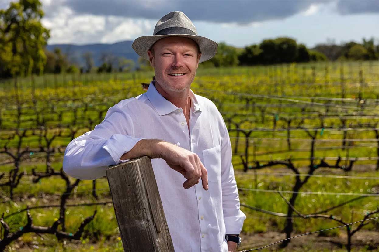 Man in a white shirt and straw hat leaning on a wooden fence post in a vineyard.